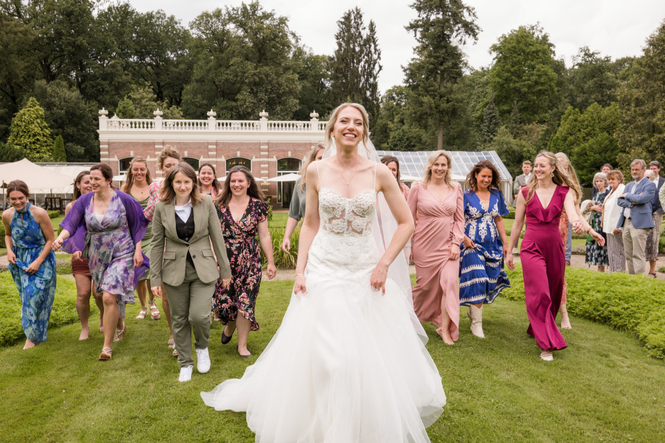 een vrouw in een trouwjurk die in het gras loopt met een groep mensen achter haar en een man in een pak en stropdas
