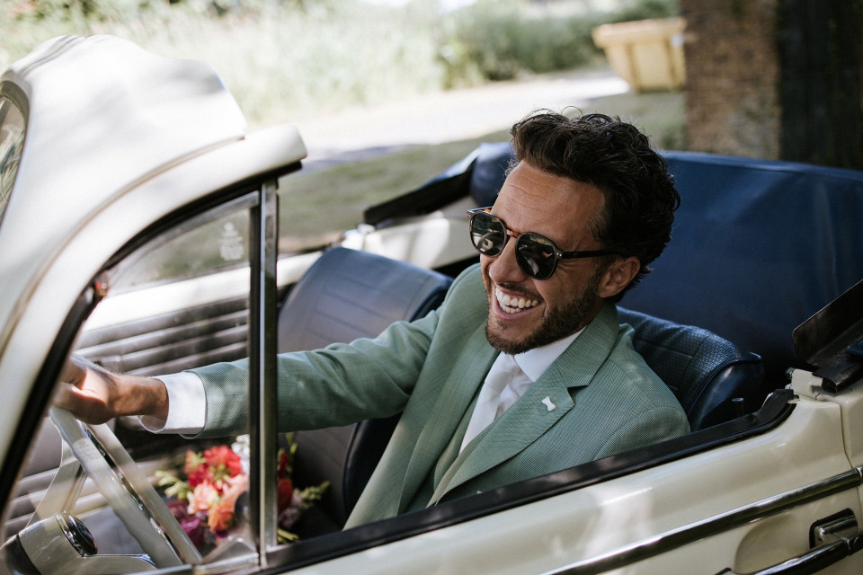 een man in een groene pak en zonnebril die een witte auto rijdt