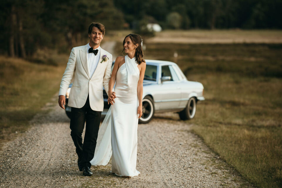 een bruid en bruidegom die een onverharde weg aflopen met een auto op de achtergrond