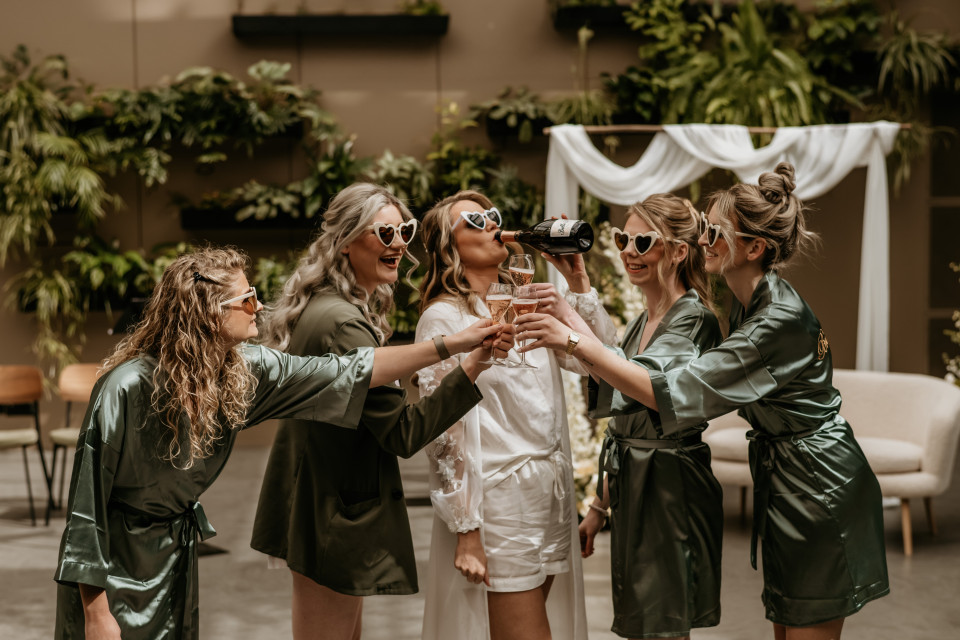 een groep vrouwen in groene gewaden die elkaar toosten in het midden van een kamer met stoelen en planten op de achtergrond