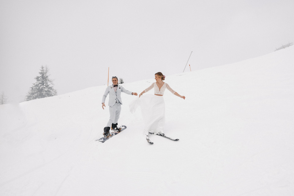 een bruid en bruidegom skiën een besneeuwde heuvel af in een witte trouwjurk en een bruidegom in een grijs pak