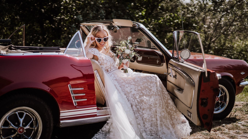 een bruid in een trouwjurk zit in de open deur van een rode cabriolet