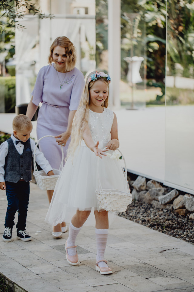 A smiling flower girl in a white tulle dress carries a basket while a young boy in a suit walks beside her at the venue.