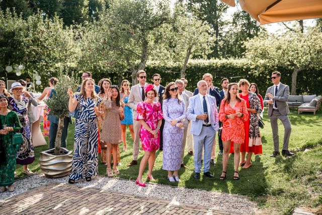 A crowd of colorfully dressed wedding guests watches and applauds in a sunlit garden.