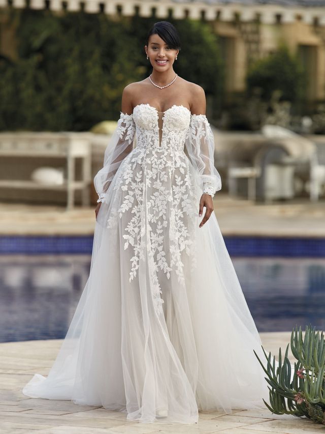 Braut in einem bestickten Brautkleid mit Off-Shoulder Ärmeln steht vor einem Pool
