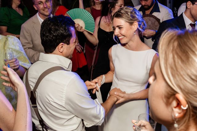 A bride and groom dance and laugh among guests on a lively party floor with colorful lighting.
