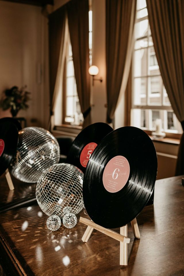 Vinyl records and disco balls used as table-number decor on wooden easels by a curtained window.