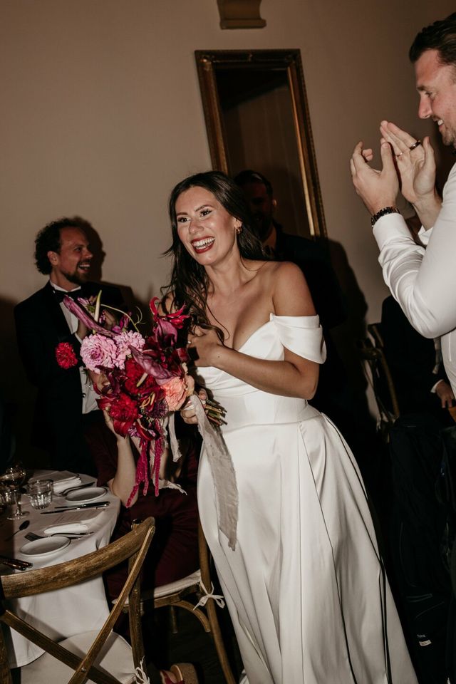 A smiling bride holds her colorful pink and red bridal bouquet while the groom applauds beside her.