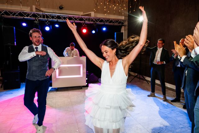 A bride throws her arms up in celebration on the dance floor as the groom and guests applaud around her.