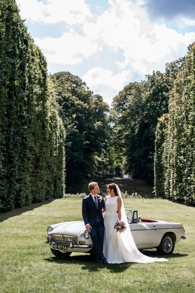 Newlyweds lean against a classic white convertible in a sunlit garden surrounded by tall trees.
