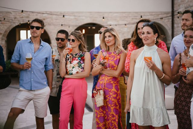 A row of colorfully dressed guests stand together with drinks watching during an outdoor reception.