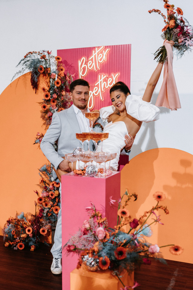 Brautpaar vor einem bunten Backdrop in orange und pink. Vor ihnen steht ein Champagner Tower. Der Backdrop ist verziert mit einem Neonschild.