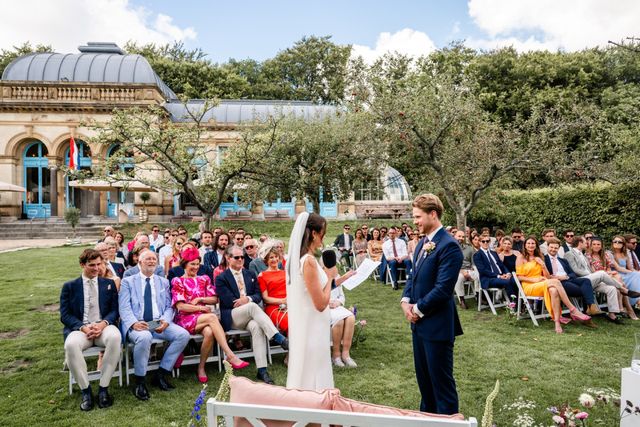 An outdoor garden ceremony with the couple standing before seated guests and an ornate orangery building behind them.