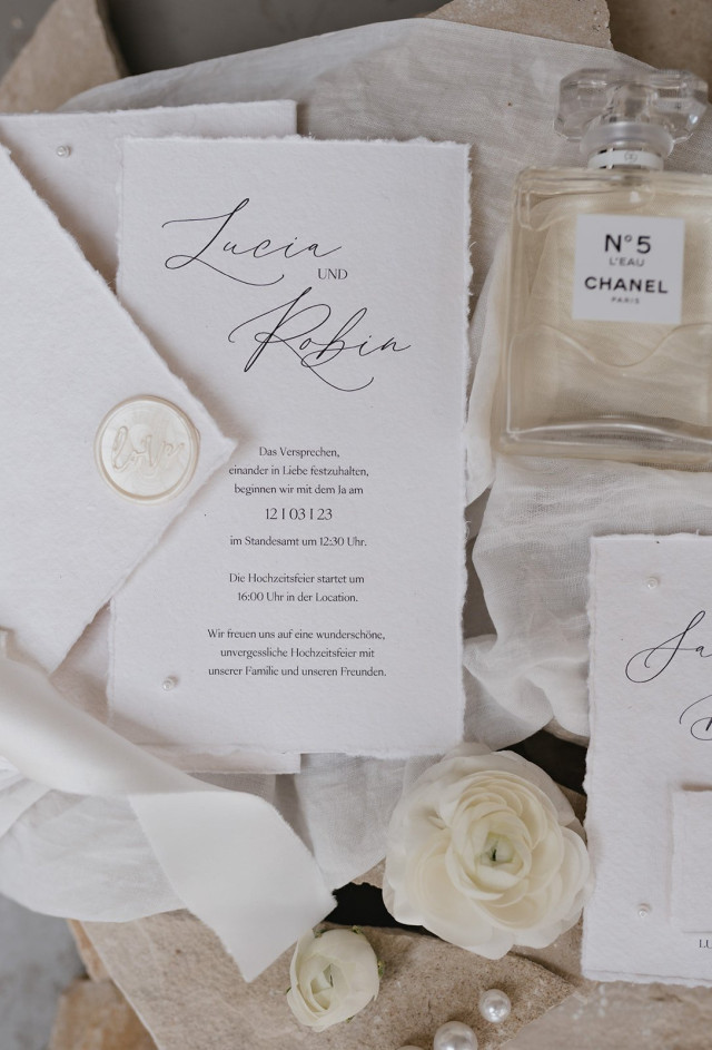 Weiße und Cremefarbene Hochzeitskarten liegen neben einer Parfümflasche