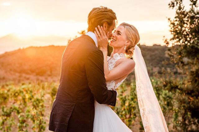A couple share a kiss at golden hour with Italian vineyard hills glowing behind them.