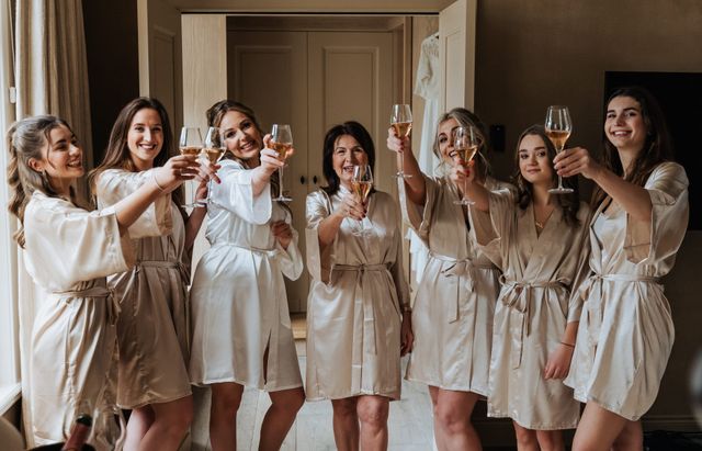 A group of women in white and beige satin robes raise champagne glasses in a cheerful toast.