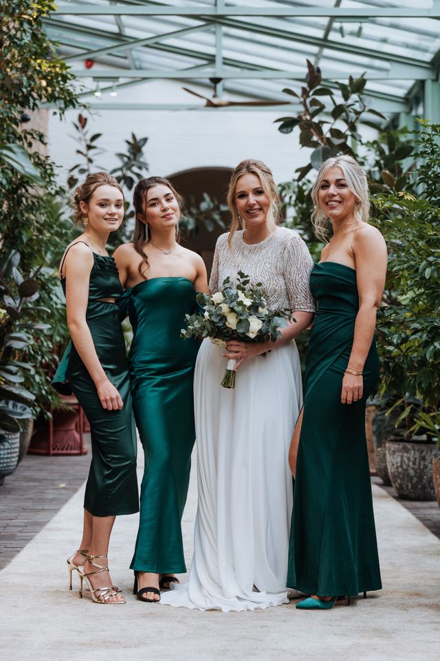 A bride poses with bridesmaids in deep emerald green satin dresses in a lush greenhouse.