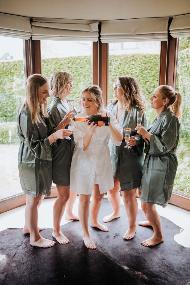 A woman in a white robe opens a gift box surrounded by friends in dark green robes by a bay window.