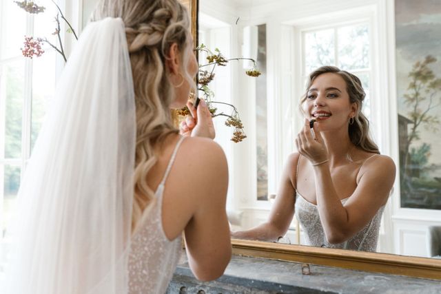 A bride touches up her lips in front of a gilded mirror, her smiling reflection visible.