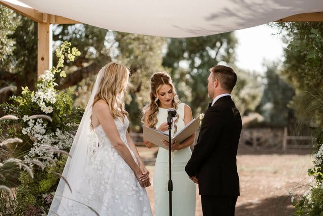 A bride, groom, and ceremony officiant stand together under a floral wedding arch.