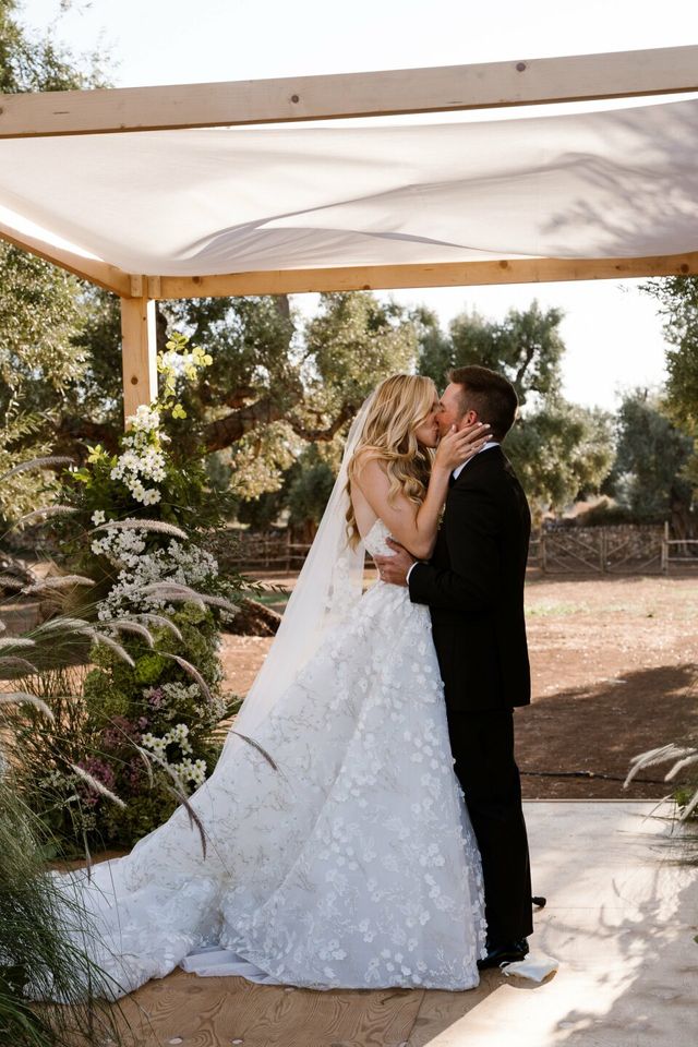A couple shares a first kiss under a wooden huppah decorated with flowers.