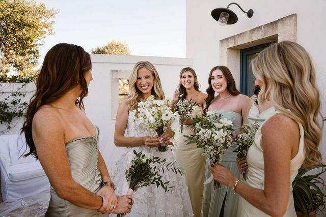A bride in a light green dress hands bouquets to her laughing bridesmaids.