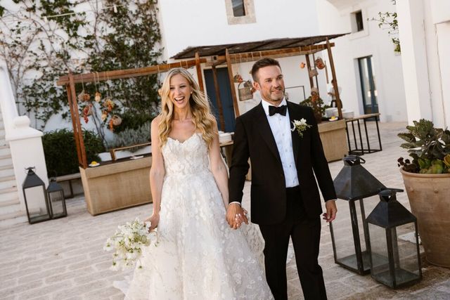 A smiling couple walks hand in hand across a terrace in front of a white building.