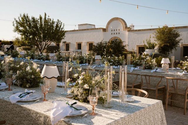 Atmospherically set outdoor tables with floral arrangements and candles before a white villa.