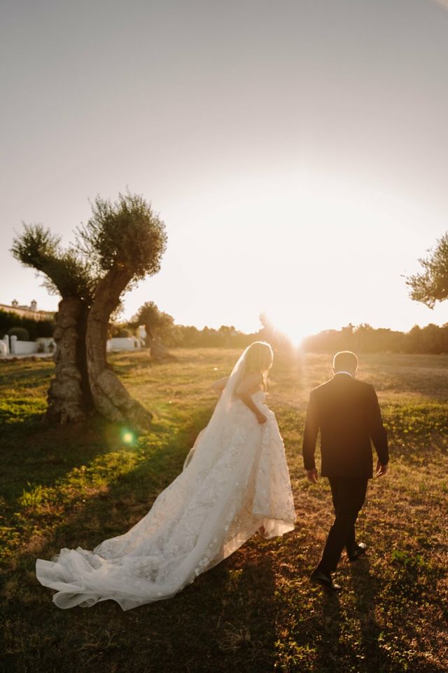 The couple walks away from the camera through a sunlit field together.
