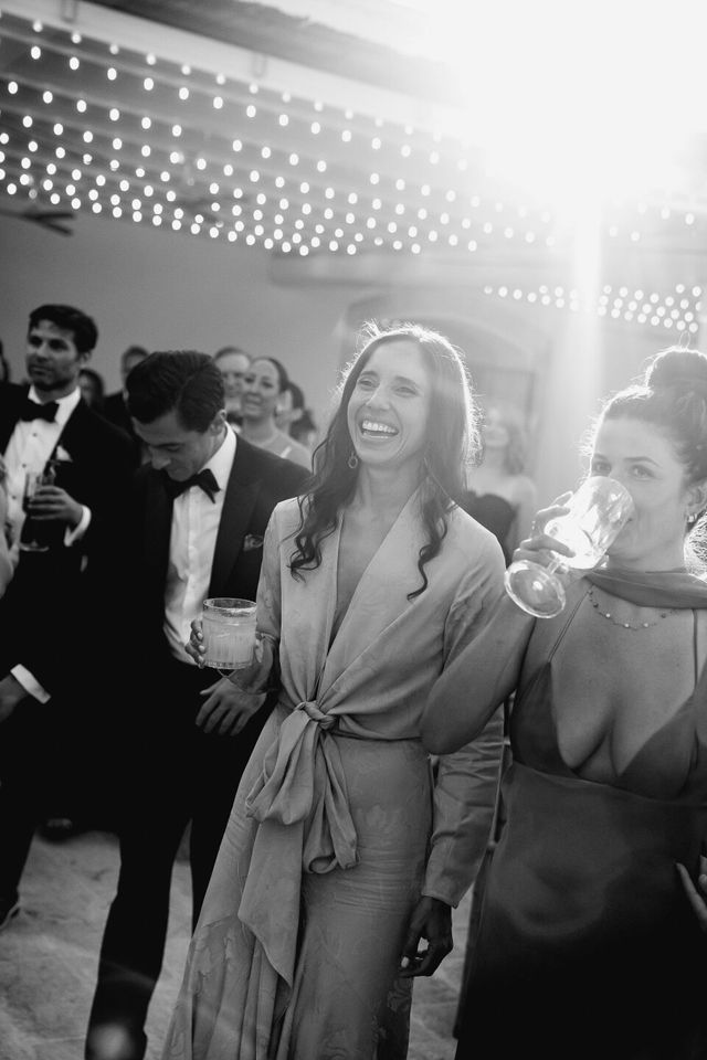 Black and white photo of laughing guests with a drink standing under string lights.
