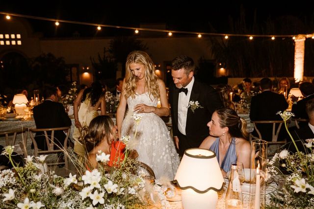 The bride and groom walk past the set dinner tables greeting seated guests in an evening ambiance with string lights.