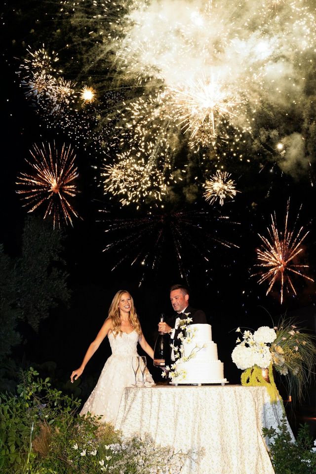 The couple stands beside their cake table with a bottle of champagne.