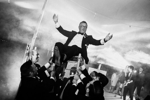 The groom is lifted into the air with arms spread by cheering guests.