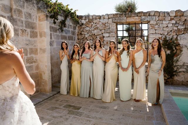 A group photo of the bride with her bridesmaids in pastel dresses beside a natural stone wall.