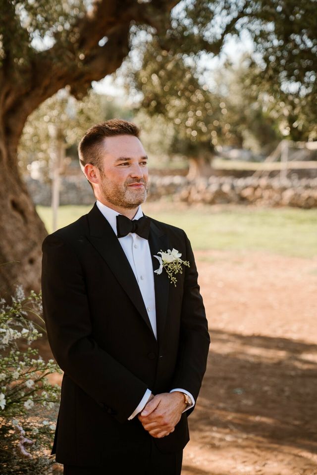 A portrait of a groom in a black tuxedo with a bow tie and boutonniere.