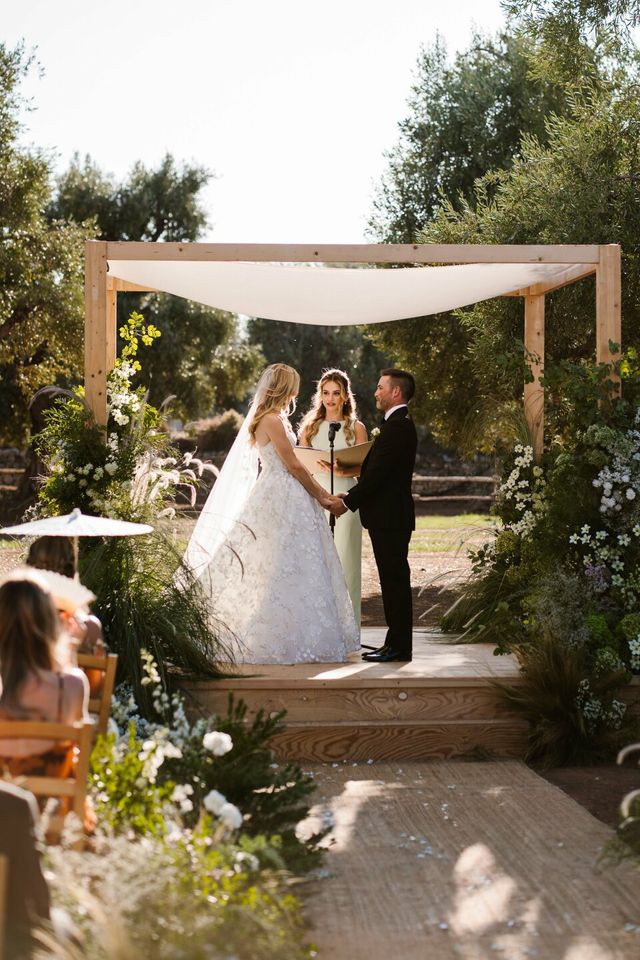 A couple holds hands under a wooden huppah during their wedding ceremony.