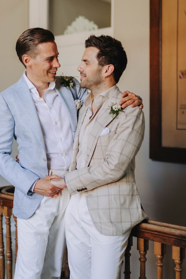 Two grooms on a staircase, one in a light blue linen suit and the other in a beige checked suit, both wearing boutonnieres.