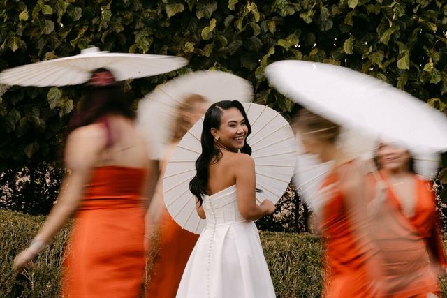 A bride holds a white parasol surrounded by bridesmaids in vibrant orange dresses, captured in motion.