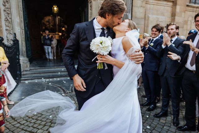 A couple kiss at the entrance of a classical building as confetti rains down and guests cheer around them.