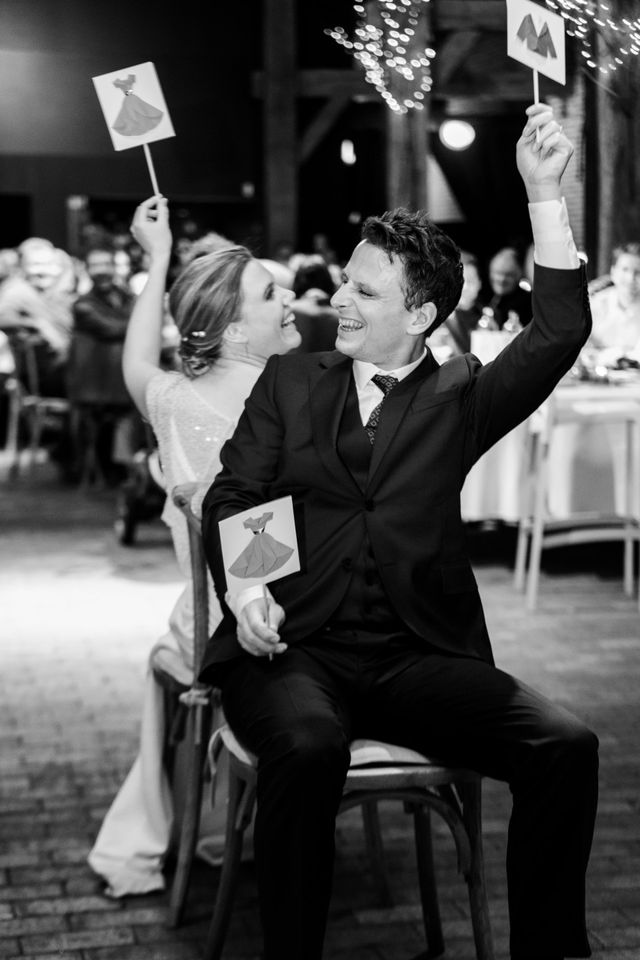 Black and white photo of a bride and groom laughing and raising cards during a fun wedding reception game.