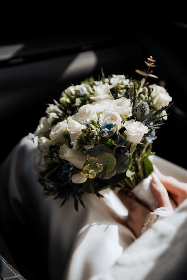 A lush bridal bouquet of white roses, blue thistles and eucalyptus in soft window light.