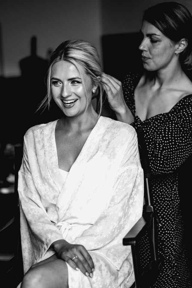 Black and white photo of a smiling bride in a white robe having her hair styled.