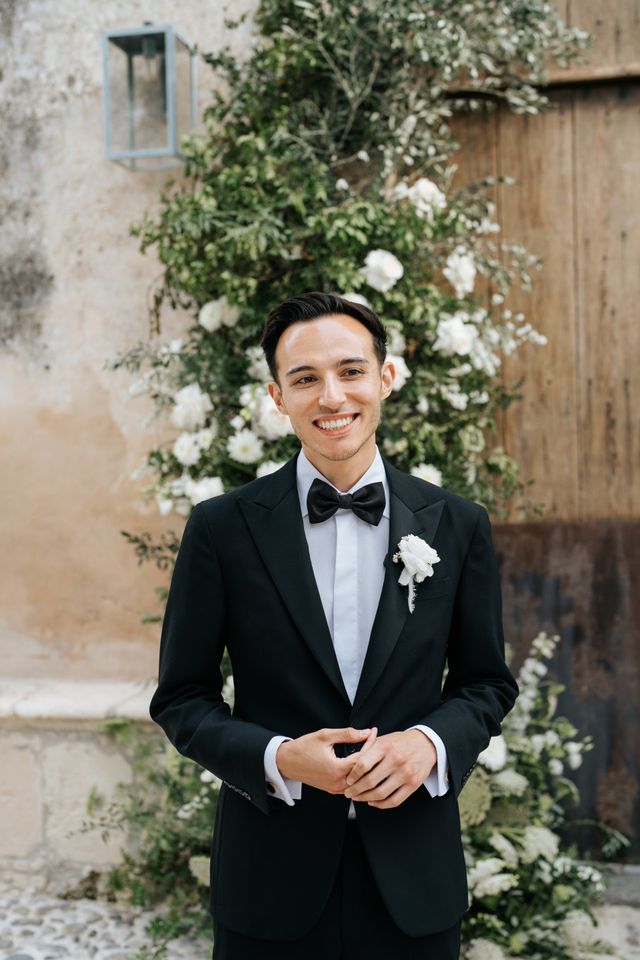 A smiling groom in a black tuxedo with a bow tie poses for a portrait.