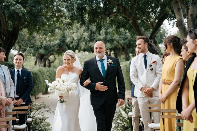 The bride is laughingly escorted by a man past guests after the ceremony, flanked by trees and onlookers.