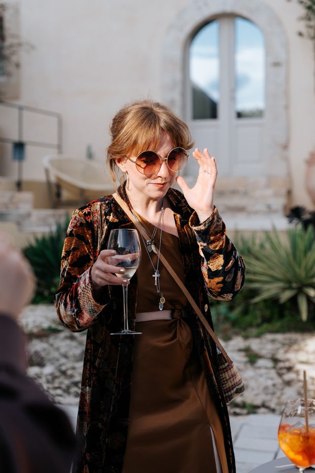 A guest in round sunglasses holds a glass of wine at the reception.