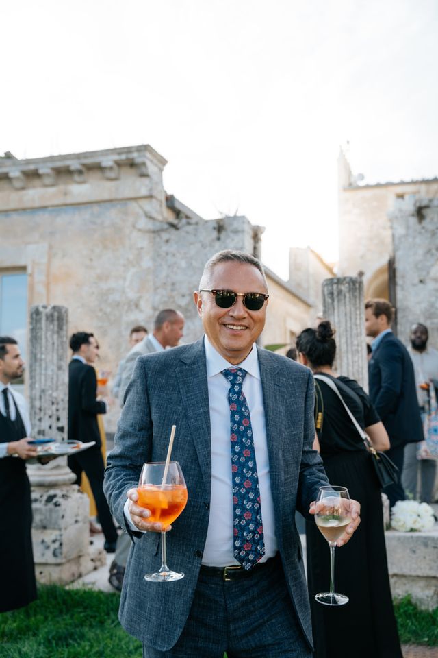 A laughing male guest in sunglasses and a grey suit holds a drink.