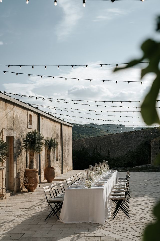 A long dinner table dressed in white linens runs across a sunny courtyard.