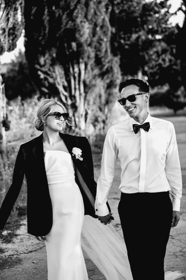 A couple in sunglasses walk hand in hand down a tree-lined lane, laughing together.