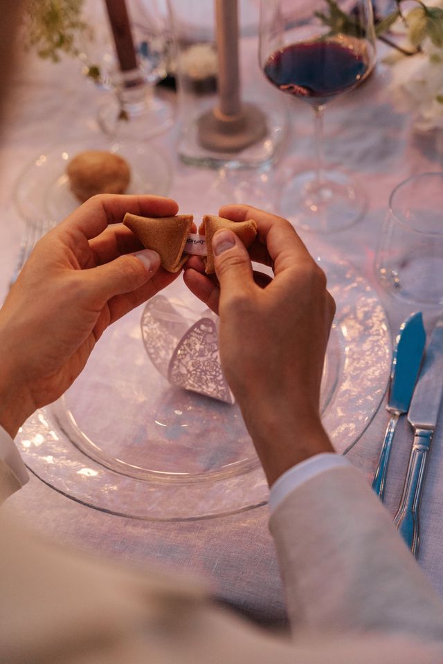 Two hands break open a paper fortune cookie above a set table with wine glasses.