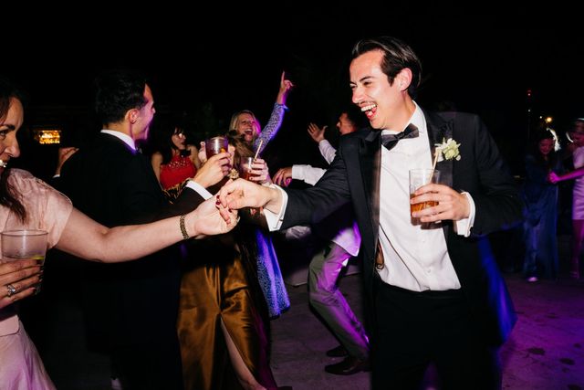 A man in a tuxedo laughs and toasts with others, drink in hand.
